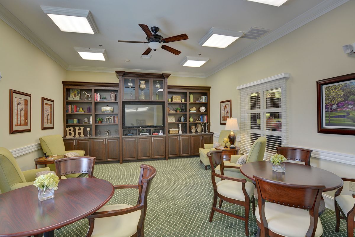 A cozy library room with bookshelves, tables, and chairs arranged neatly on a carpeted floor.