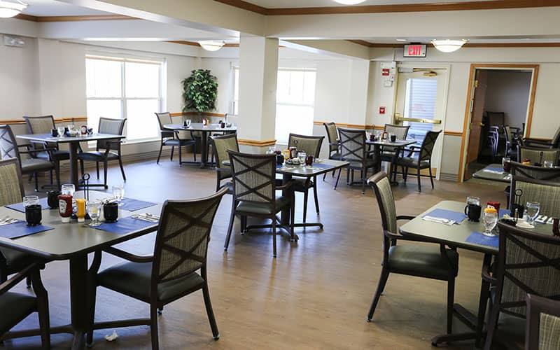 An empty dining area with neatly arranged tables and chairs, illuminated by natural light through large windows.