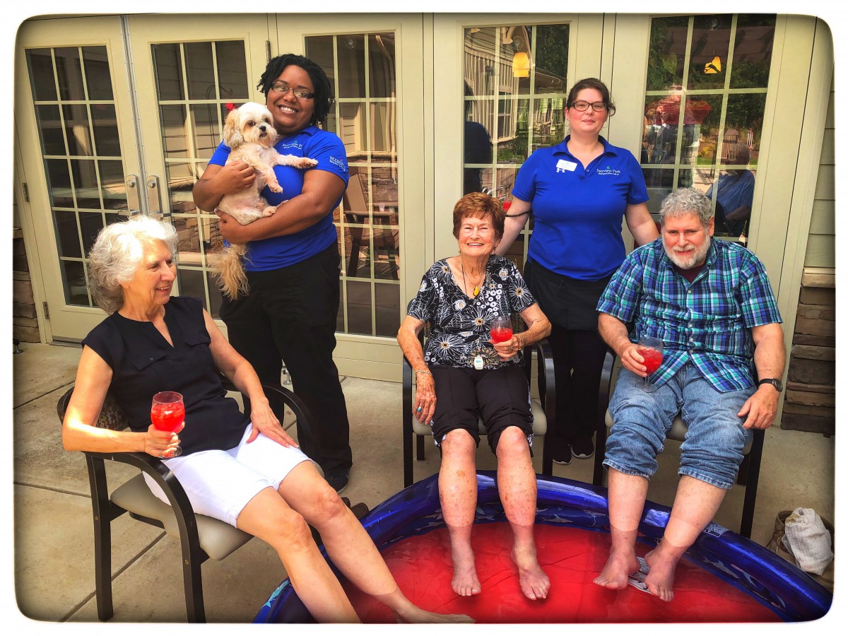 A group of people, including seniors, relaxing outdoors with drinks and a small pool.