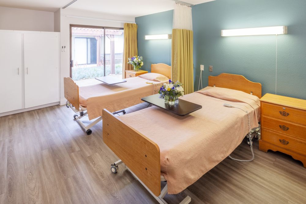 A clean and well-lit hospital room with two patient beds and bedside tables.