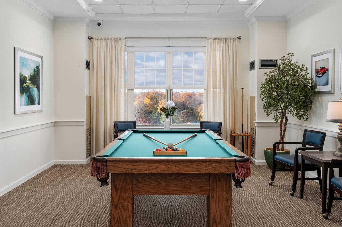 A well-lit game room featuring a classic pool table, set against large windows with curtains and surrounded by cues and comfortable seating.