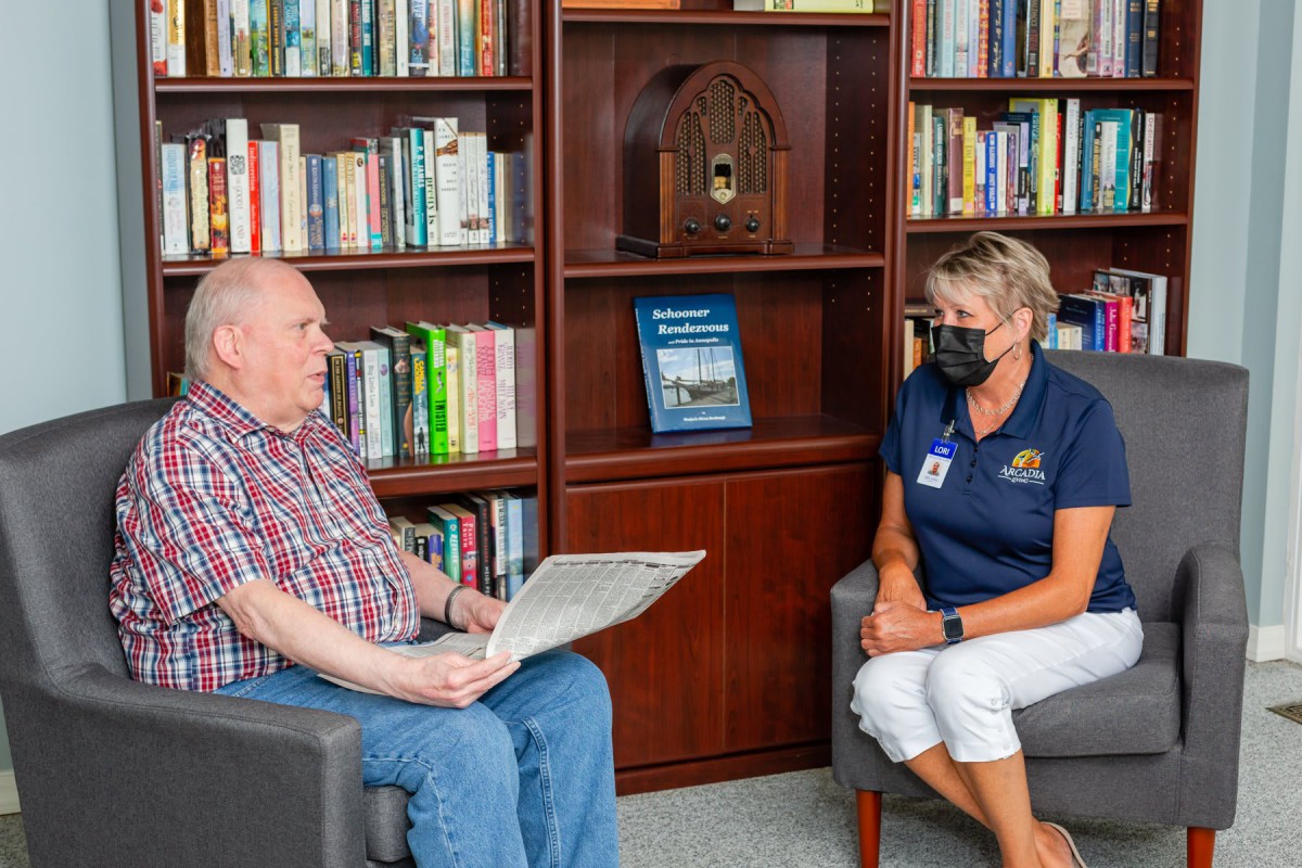 Two people sitting in a library engaged in conversation, with one person holding a newspaper.