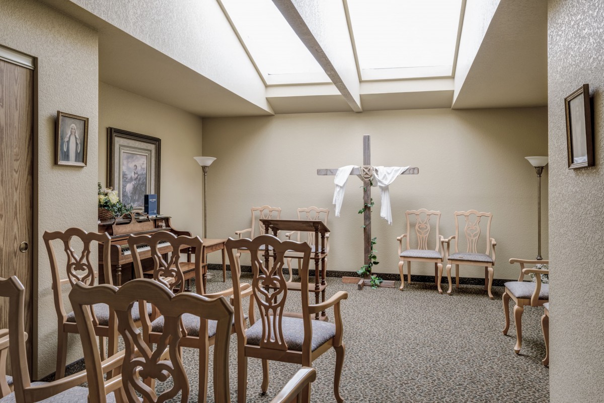 A small chapel interior with arranged chairs, a cross adorned with white drapery, and natural light streaming through skylights.