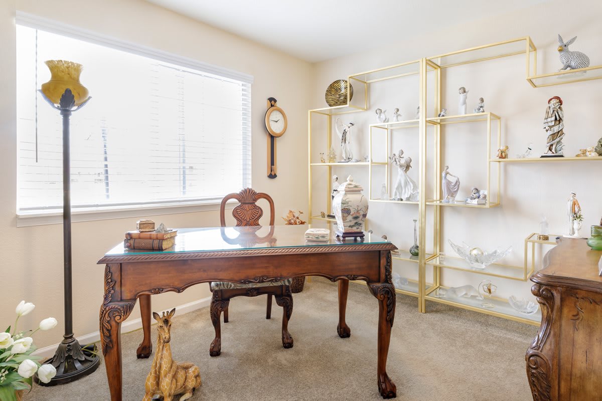 A cozy home office with a classic wooden desk, elegant shelves, and various decorative items bathed in natural light.