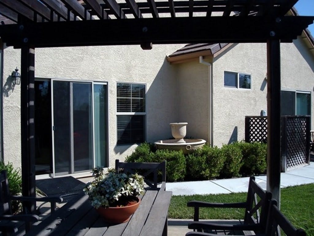 A cozy backyard patio with a pergola, table, chairs, and potted plants next to a house with a sliding glass door.