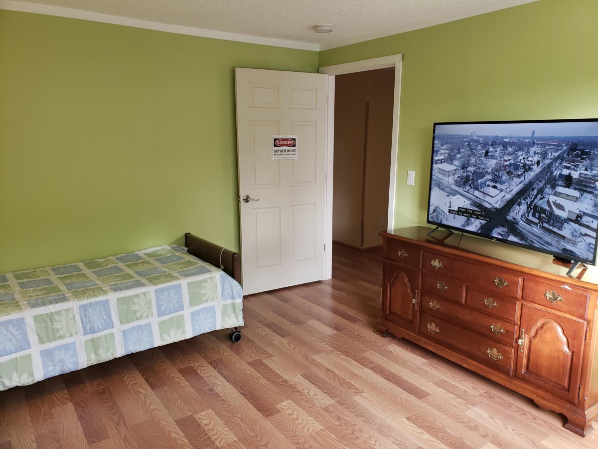 A bedroom with green walls, featuring a bed, a wooden dresser with a television on top, and hardwood flooring.