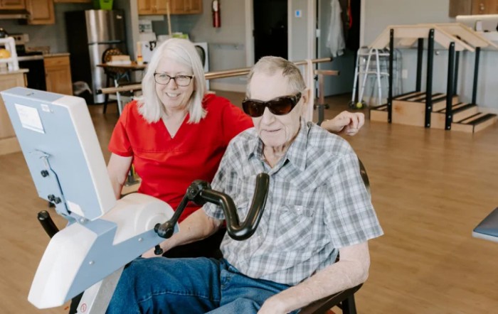 An elderly couple, with the man in a wheelchair, sits indoors smiling at the camera. He is wearing sunglasses.