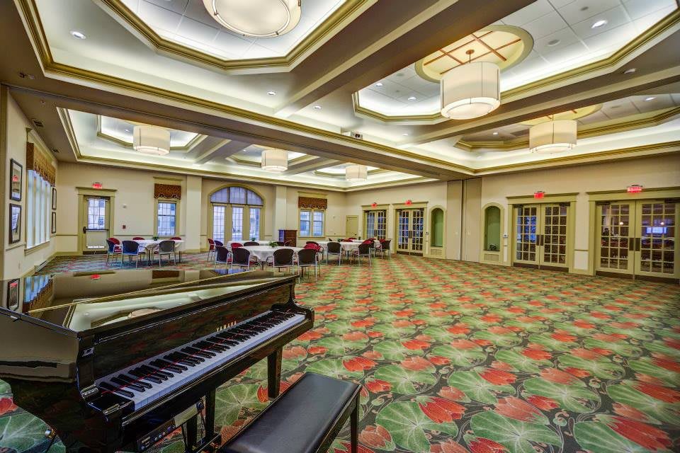 A spacious conference room with a grand piano, carpeted floors, and elegant ceiling lighting.