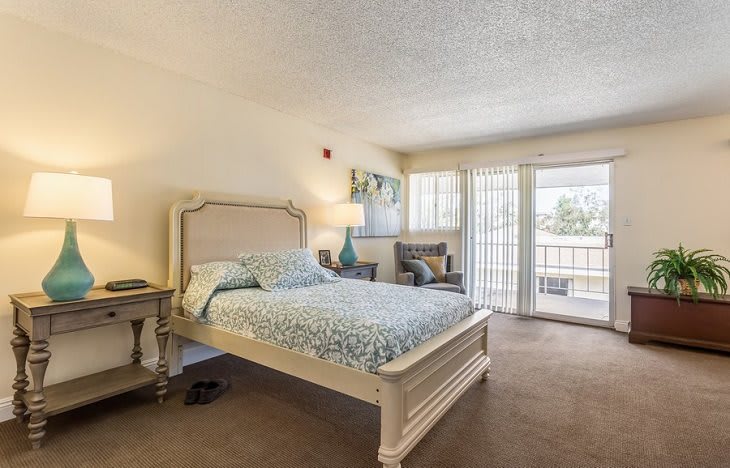 A cozy, well-lit bedroom with a bed, lamp, and chair near the window.