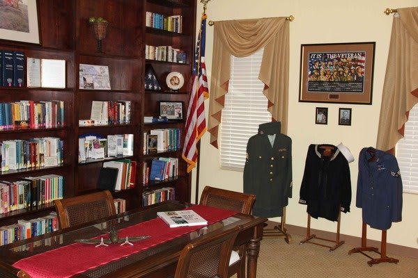 A cozy office library featuring a bookshelf, military uniforms on display, and an American flag by the window.