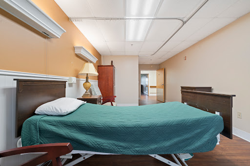 A well-lit hospital room featuring a bed with a green blanket and a bedside lamp.