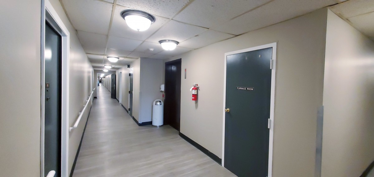 A hallway with several doors, ceiling lights, a fire extinguisher, and a trash can.