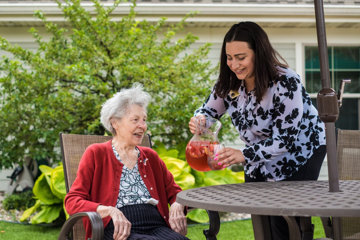 A young woman serves a drink to an elderly woman in a garden setting.