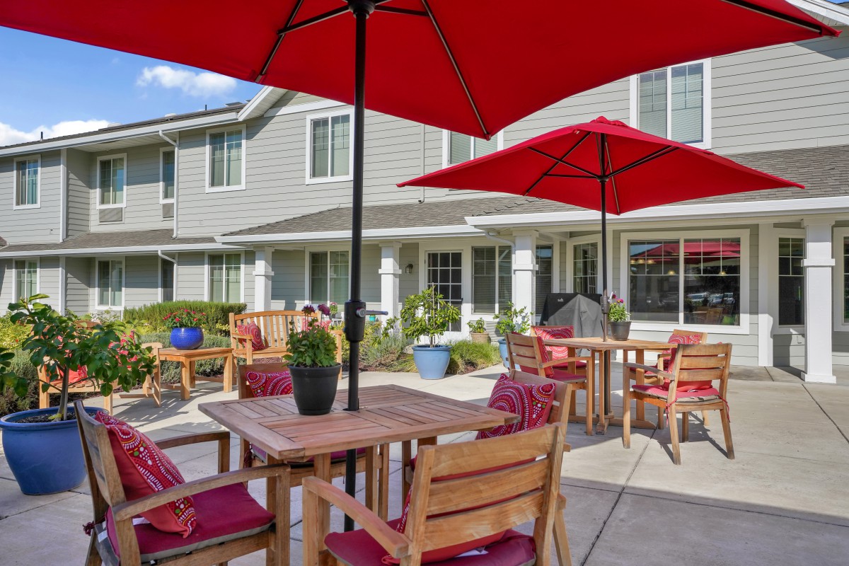 A cozy outdoor patio with wooden furniture and red umbrellas, set against a building backdrop.