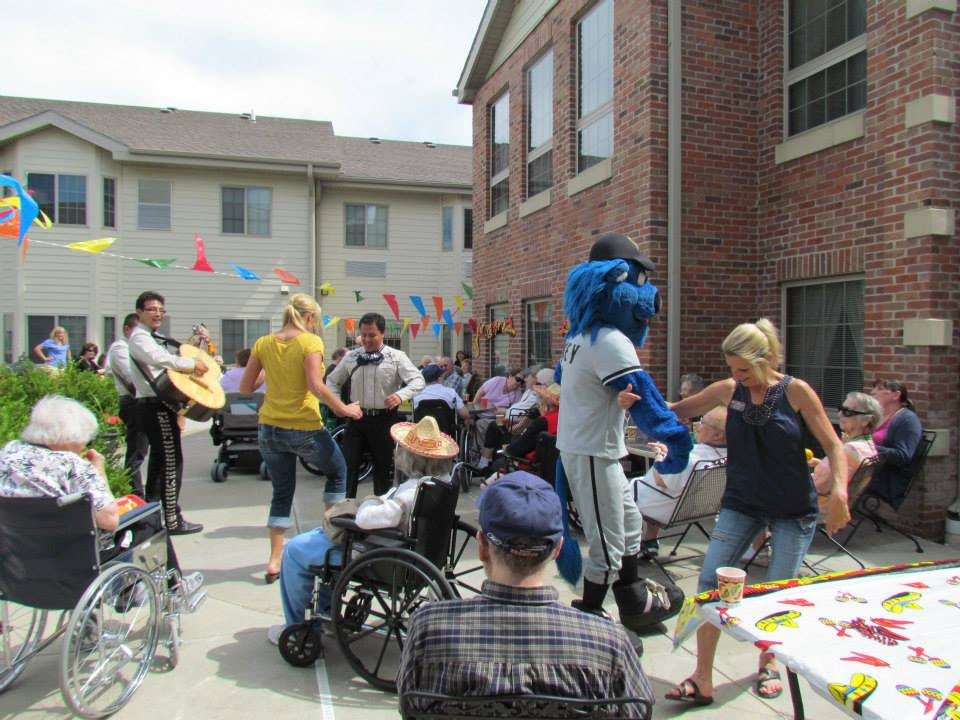 A lively outdoor event with people gathered, including a mascot in costume and individuals in wheelchairs, surrounded by festive decorations.