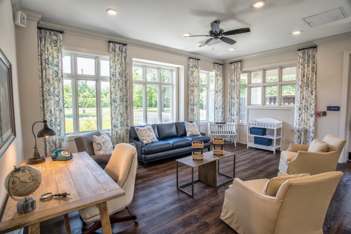 A cozy and well-lit living room featuring large windows, a sofa, chairs, and wooden flooring.