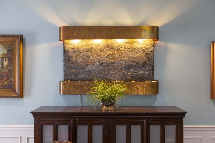 A stylish indoor wall fountain with decorative lighting and a potted plant placed on a wooden console table.