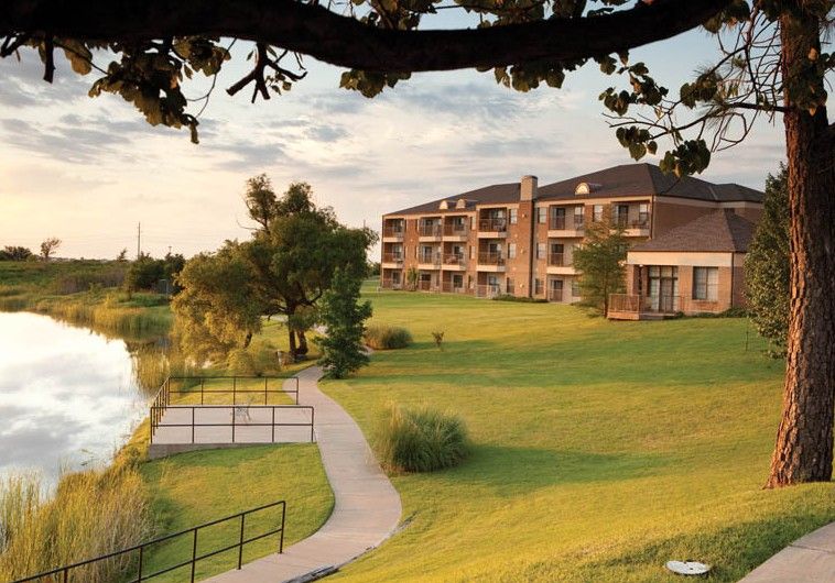 A scenic view of a hotel by the lake surrounded by lush greenery and a tranquil pathway during sunset.