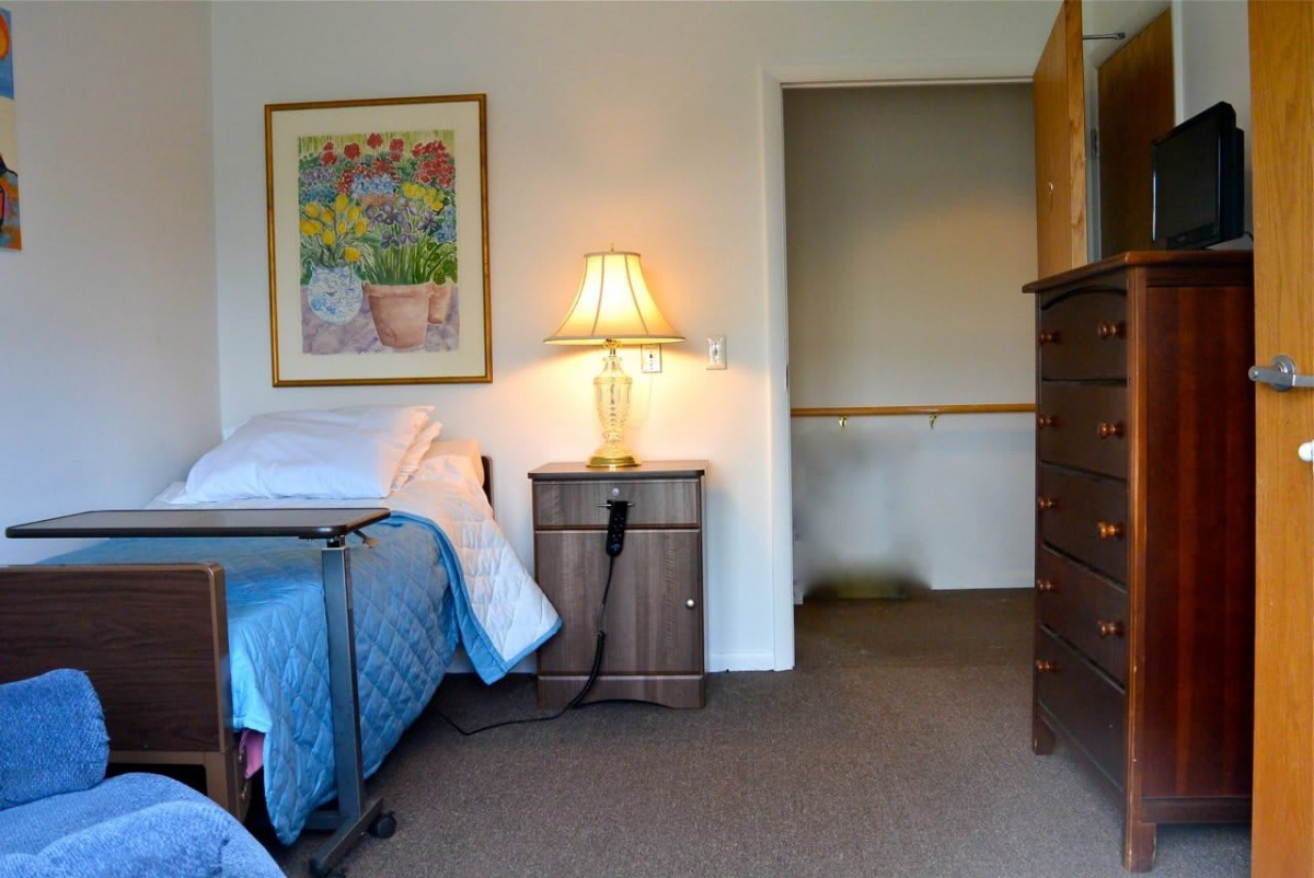 A cozy bedroom featuring a neatly made bed, a wooden dresser, and a bright lamp on the nightstand.