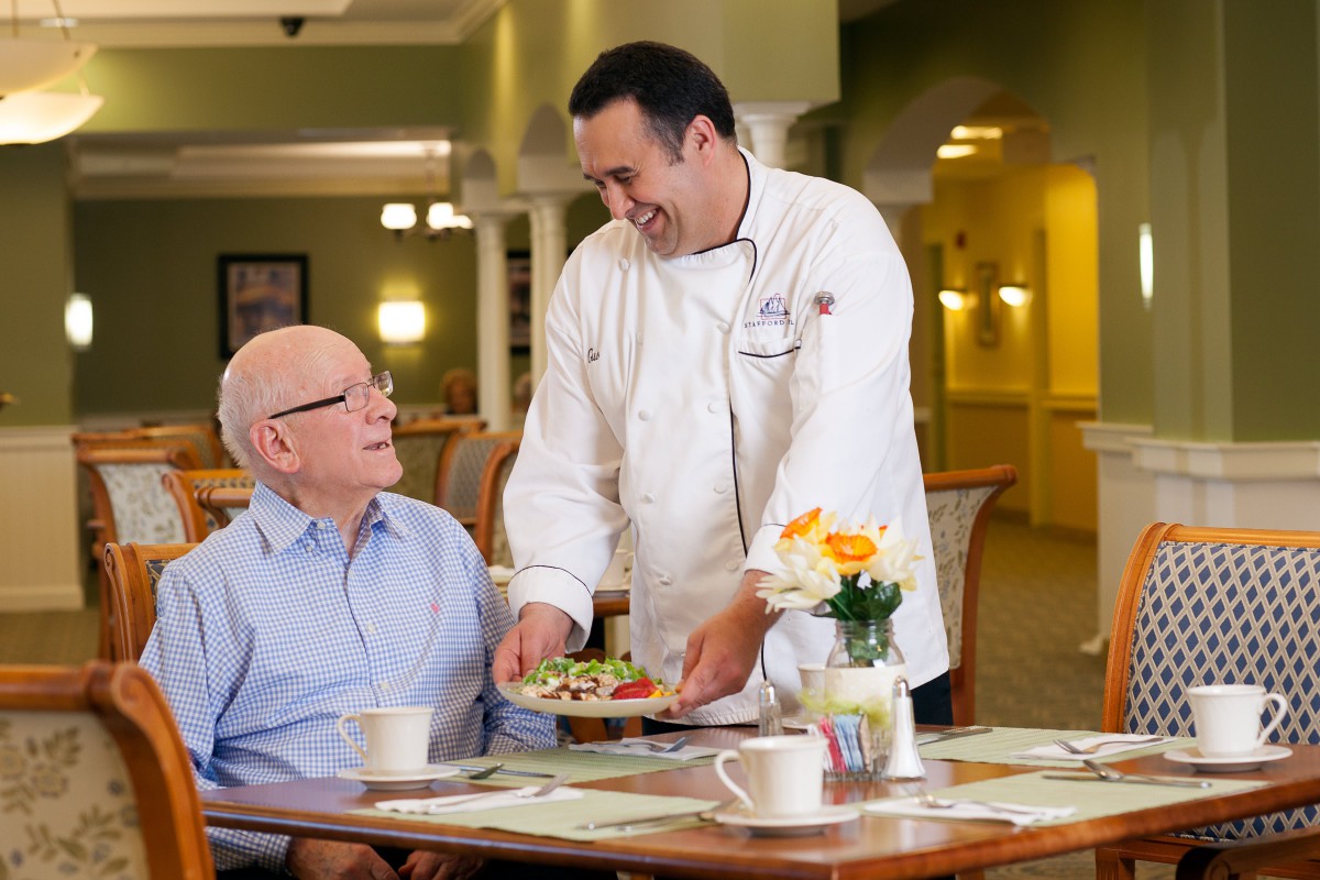 A chef serving a meal to a senior man in a dining room setting.
