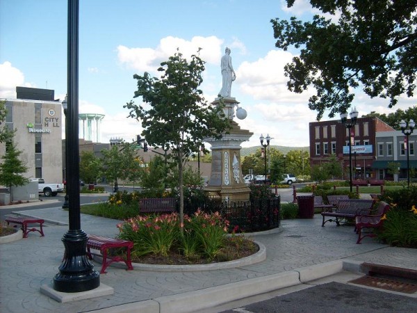 A small urban park featuring a statue surrounded by benches, trees, and landscaping.