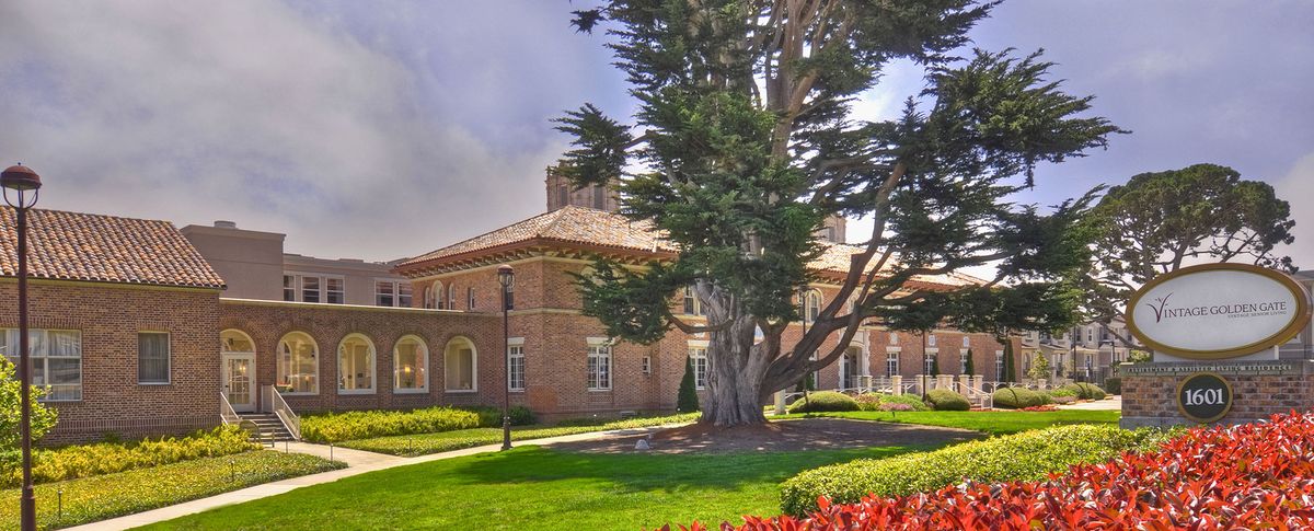 A brick building surrounded by lush greenery and a large tree in a landscaped garden.