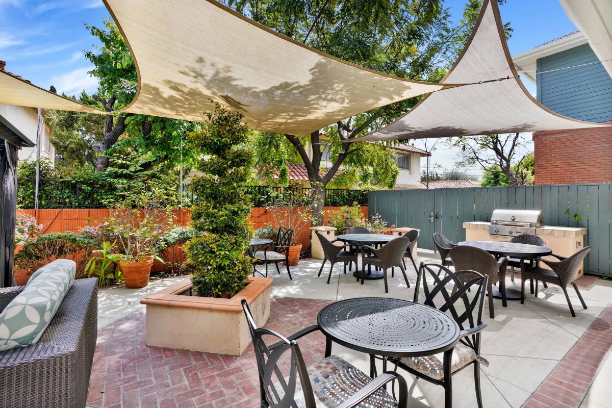A cozy outdoor patio with tables and chairs under shade sails, surrounded by lush greenery.
