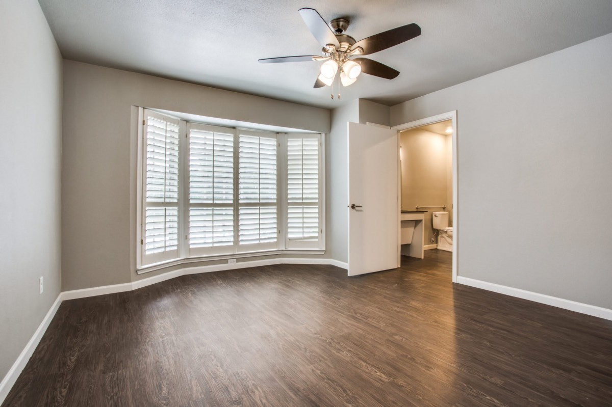 A room with hardwood floors, a ceiling fan, and window shutters, featuring a doorway leading to a bathroom.
