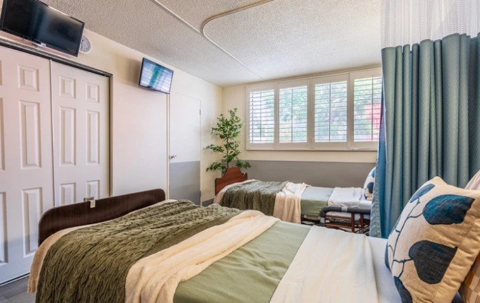 A well-lit hospital room with two neatly made beds, a wall-mounted television, and a large window with shutters.