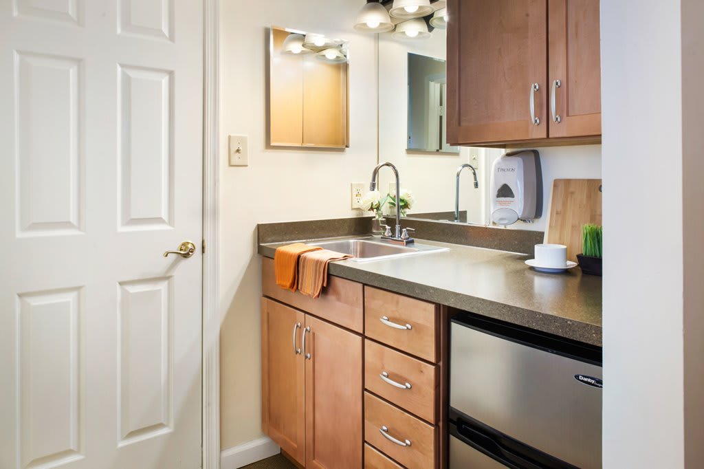 A compact kitchenette with wooden cabinets and a sleek countertop, featuring a sink and mirror.
