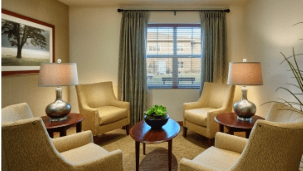 A cozy living room with four beige armchairs arranged around a wooden table, flanked by two lamps and set against a window with curtains.