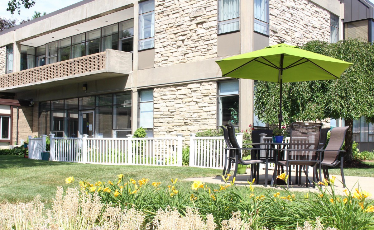 A modern building with a patio area featuring a table and chairs under a green umbrella, surrounded by a garden with flowers.