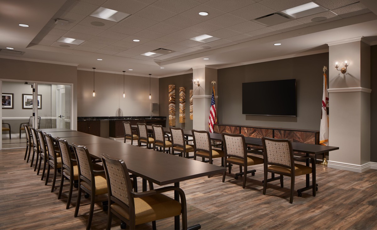 A well-lit conference room featuring a long table, multiple chairs, a large television on the wall, and an American flag in the corner.