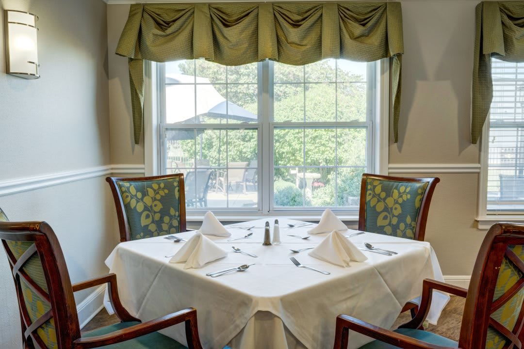 A neatly set dining table near a window with elegant curtains, surrounded by chairs.