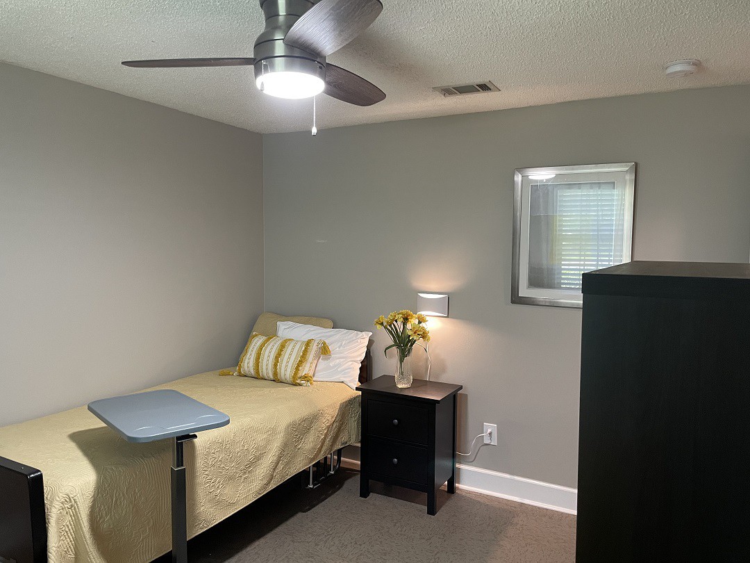 A small, modern bedroom with a single bed, nightstand, lamp, and ceiling fan.