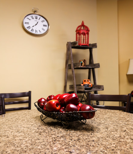A cozy kitchen scene featuring a bowl of red apples on a granite table, complemented by a wall clock and decorative shelves.