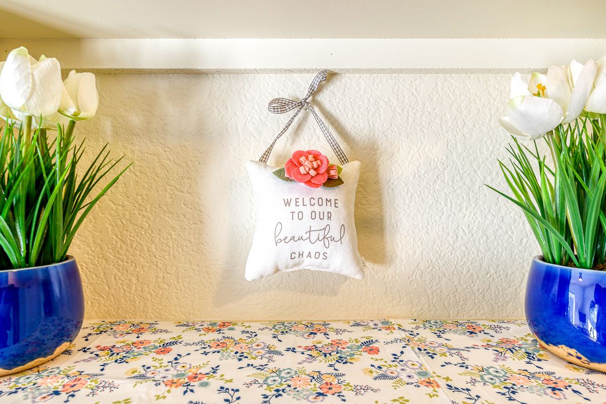A small cushion with a 'Welcome to Our Comfortable Home' message hangs on the wall above a floral patterned surface.