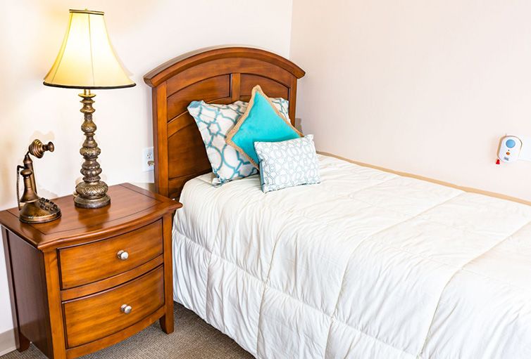 A cozy bedroom with a single bed, featuring neatly arranged pillows and a wooden nightstand with a lamp.