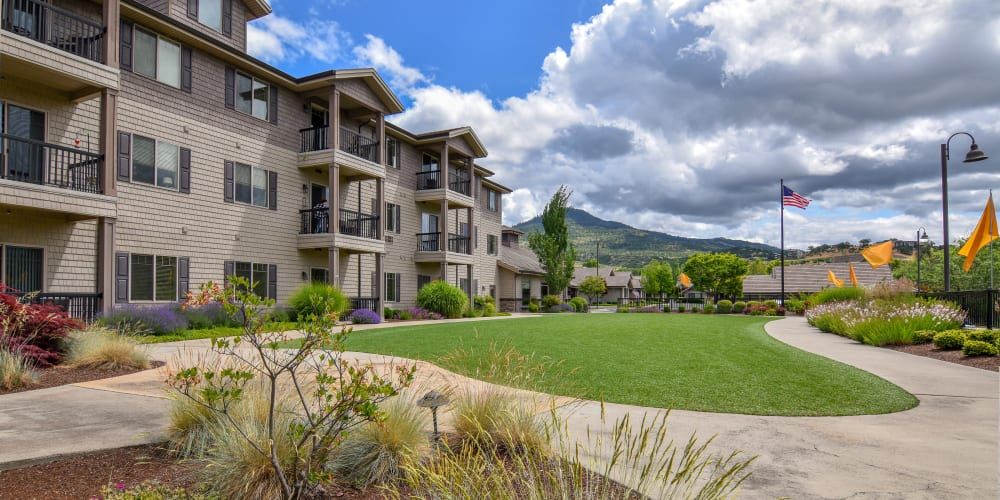 A modern apartment building with balconies set against a backdrop of mountains and a cloudy sky, surrounded by green lawns and landscaped gardens.