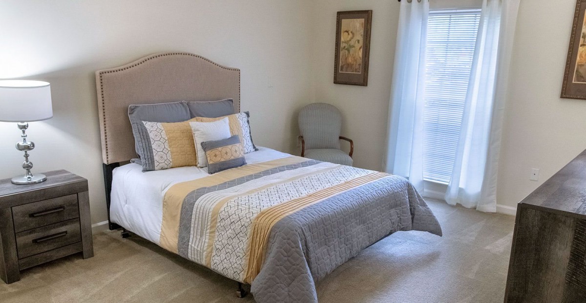 A neatly decorated bedroom with a bed, upholstered chair, artwork on the wall, and light coming through a window.