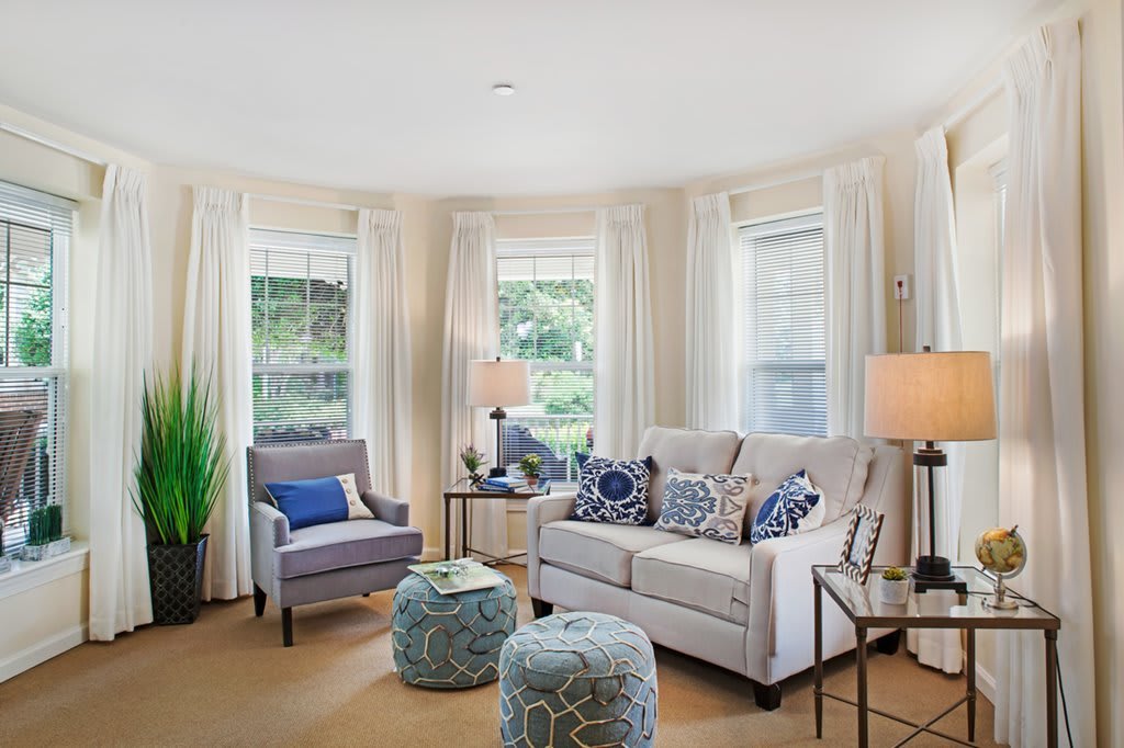 A cozy living room with a comfortable sofa and armchair, adorned with blue pillows and surrounded by large windows with curtains.