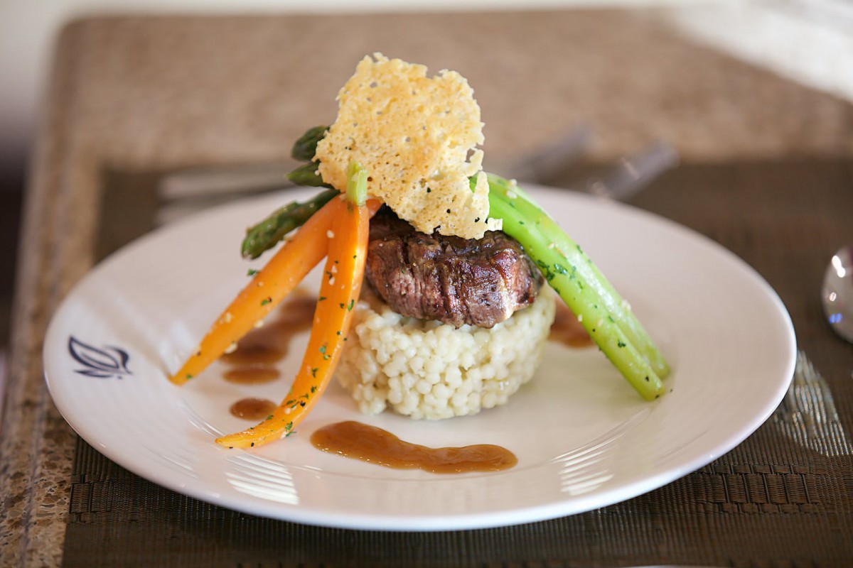 A gourmet dish featuring steak on a bed of risotto, garnished with asparagus, carrots, and a parmesan crisp.
