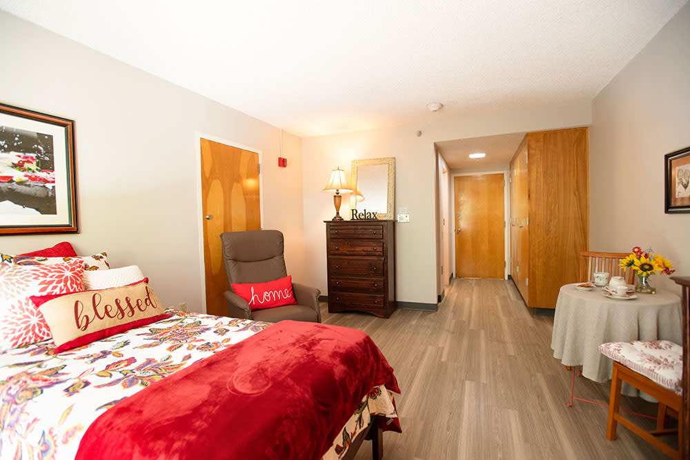 A cozy bedroom featuring a bed with a red blanket, an armchair, and wooden furniture on a wooden floor.
