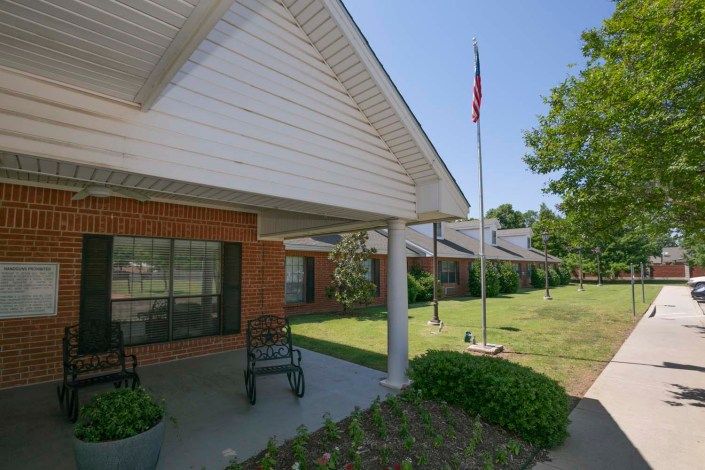 A porch with rocking chairs and a flagpole outside a brick house on a sunny day.