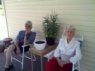 An elderly couple sitting on a porch, enjoying a relaxed moment together with potted plants nearby.
