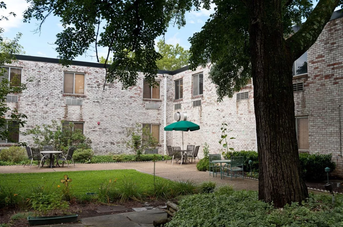 A serene courtyard with a stone building backdrop, shaded by trees and featuring outdoor seating with an umbrella.