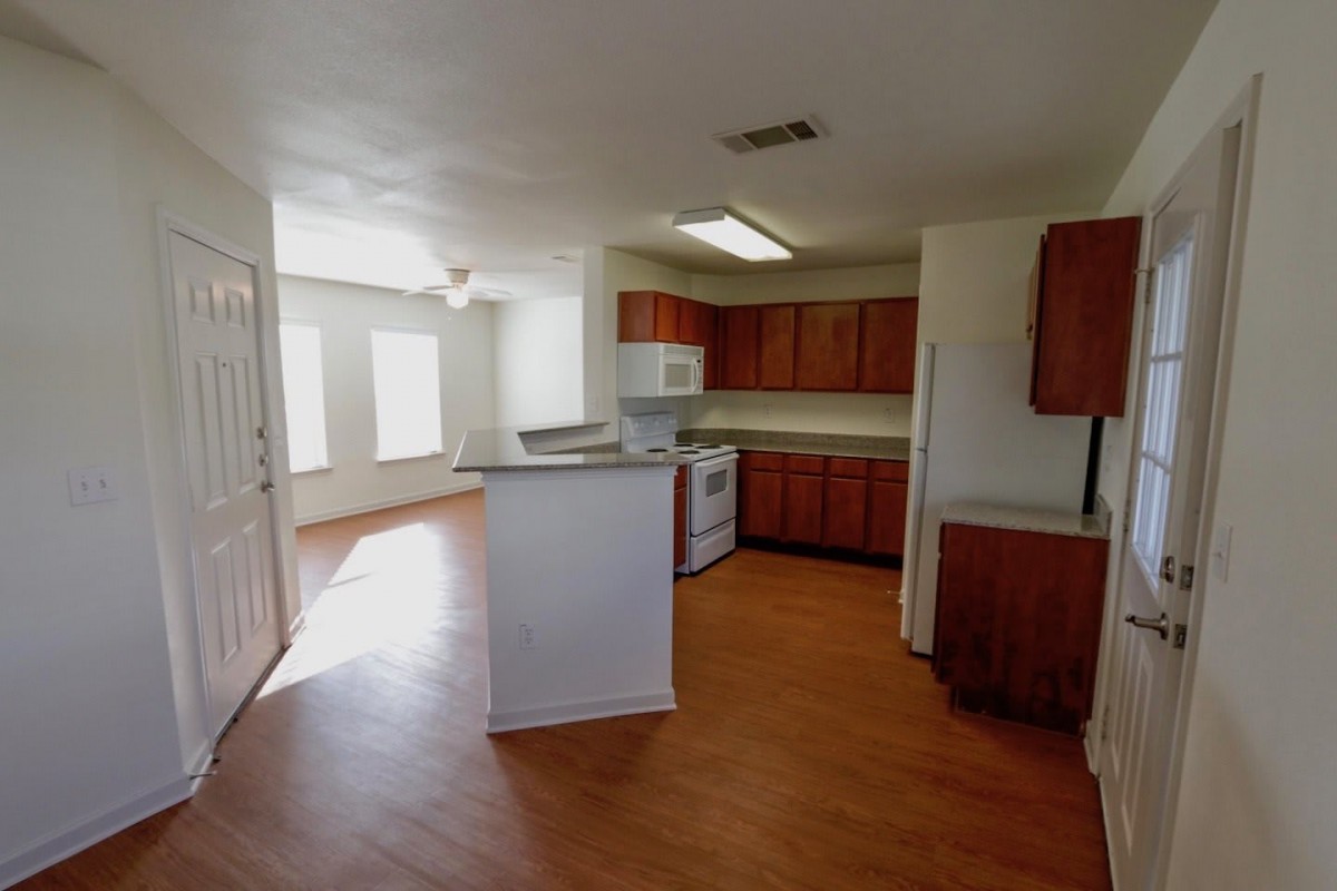 A bright kitchen with wooden cabinets and white appliances, featuring an open layout with ample natural light on a hardwood floor.