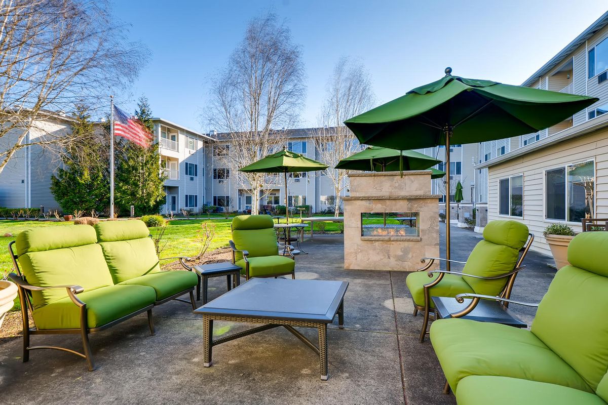 A modern outdoor seating area with green cushioned chairs and tables, complete with umbrellas and a fireplace at an apartment complex.