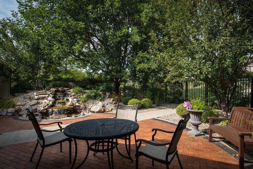 A serene patio setup with a round table and chairs, surrounded by lush greenery and a peaceful garden landscape.
