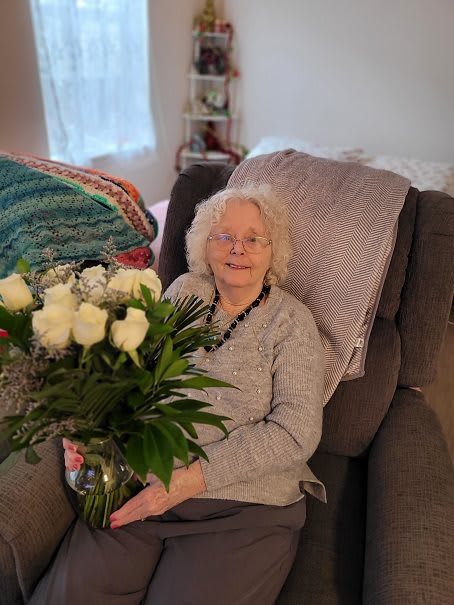 An elderly woman sitting on a sofa, holding a bouquet of white roses and smiling.
